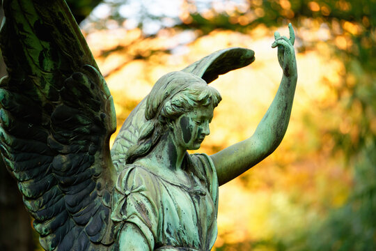 an angel with wings in a cemetery in front of luminous autumn leaves in the background raises one arm to the sky