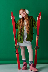 excited preteen girl in ear muffs and winter attire holding red skis on turquoise background