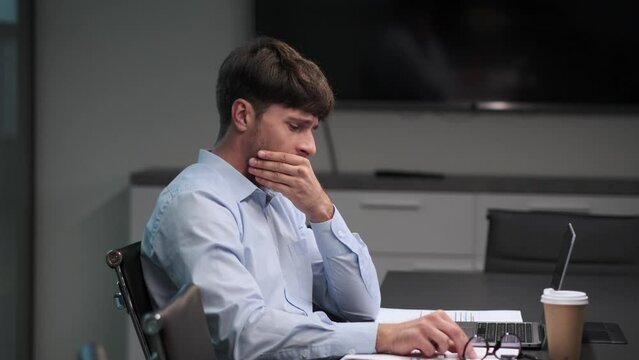 In the midst of a workday, a tired businessman lets out a yawn at desk, the strain of exhaustion evident in eyes. The office is filled with a sense of weariness as he battles fatigue to complete task.