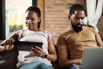 African American couple using digital tablet and laptop while relaxing on sofa in the living room