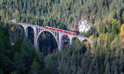 A red passenger train is crossing the famous Wiesener viaduct on the train line Davos - Filisur, the highest viaduct in swiss alps - obrazy, fototapety, plakaty