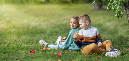 A little girl and boy are sitting on the green grass in the garden. They are holding an apple pie in their hands and smiling. Red apples lie next to them. Friendship and happy family concept.