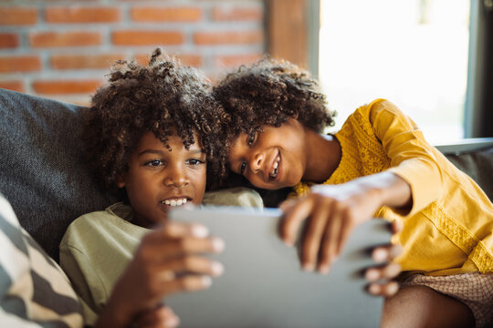 Little African American kids relaxing on sofa at home and watching something on digital tablet