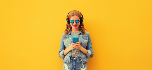 Portrait of stylish modern happy young woman listening to music in headphones with phone wearing jacket, sunglasses on yellow background