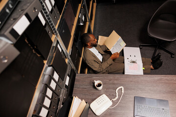 Detective sitting on office floor searching csi report in folder at night time. Exhausted investigator studying crime case files while working on investigation overtime top view