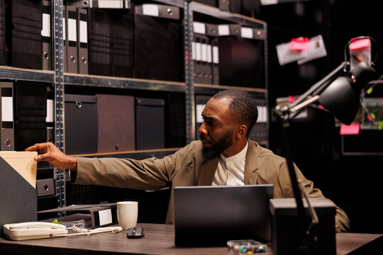 Detective analyzing crime case and searching police report while conducting investigation. African american policeman working in office and studying archival records in evening