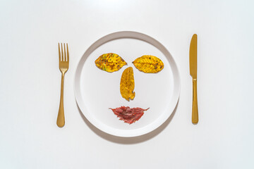 A white plate with three autumn leaves and a red leaf arranged in a smiley face. There is a gold fork on the left side of the plate and a gold knife on the right side. Concept of art and creativity