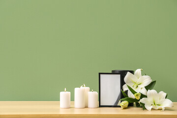 Blank funeral frame, burning candles and beautiful lily flowers on wooden table near color wall