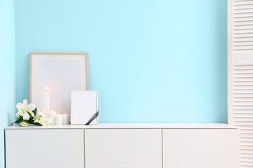Blank funeral frame, beautiful lily flowers and burning candles on white cabinet near color wall