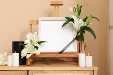 Blank funeral frame with lily flowers, mortuary urn and burning candles on wooden cabinet against...