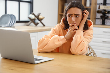 Young afraid woman with laptop sitting at table in kitchen
