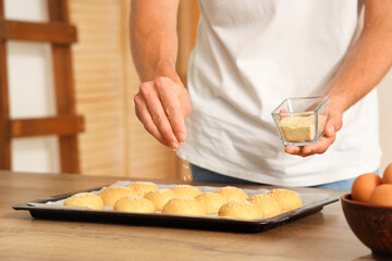Handsome young man sprinkling raw buns with sesame seeds at table in bakery