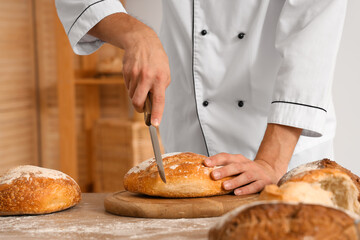 Handsome young chef cutting fresh bread on table in bakery, closeup