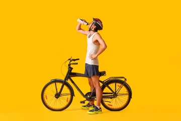 Poster Young male cyclist with bicycle drinking water on yellow background © Pixel-Shot