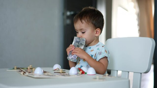 Adorable kid sitting at desk drinks juice from doypack. Toddler looks focused on the toys in front of him.