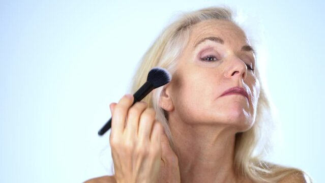 Closeup portrait of 50s, 60s, mature middle aged nude woman applying natural concealer makeup powder foundation with large brush on clean face skin smoothing wrinkles, white background.