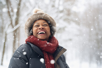A senior african american woman is playing in the snow happily with a winter coat and a winter hat in a in snow covered forest during day in winter while snowing - Powered by Adobe
