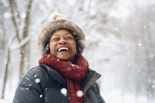 A senior african american woman is playing in the snow happily with a winter coat and a winter hat in a in snow covered forest during day in winter while snowing