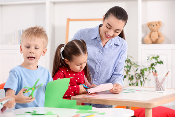 Fototapeta premium Nursery teacher and cute little children making toys from color paper at desks in kindergarten. Playtime activities