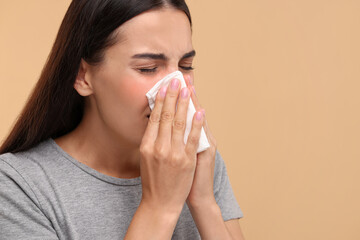 Suffering from allergy. Young woman blowing her nose in tissue on beige background, closeup. Space for text