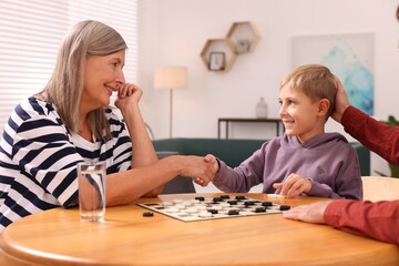 Family playing checkers in room. Grandmother shaking hands with her nephew after game - obrazy, fototapety, plakaty