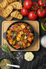 Dish with tasty ratatouille, ingredients and bread on black table, flat lay