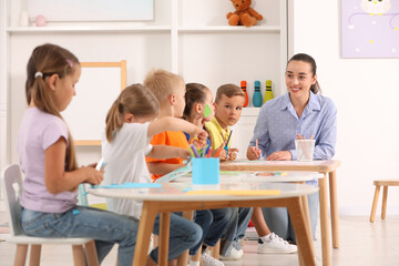 Nursery teacher with group of cute little children drawing and cutting paper at desks in...