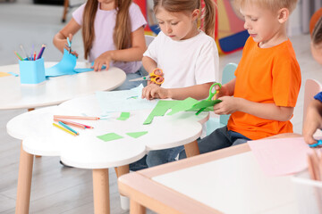 Cute little children cutting color paper with scissors at desks in kindergarten. Playtime activities