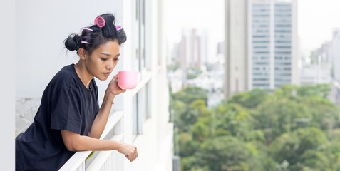 A woman in curlers holds a mug on the balcony of an apartment building in a residential area