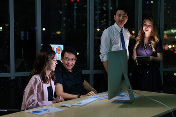 Asian professional skilled male programmer helping fixing and explaining computer problems for businesswoman employee working night shift overtime with male female businessman colleagues in office