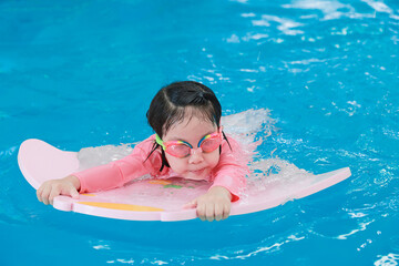 Smiling little girl learning to swim in pool with flutterboard during swimming class