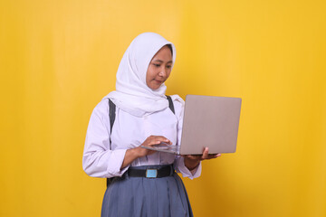 Young Asian muslim female student in school uniform using laptop isolated on yellow background - obrazy, fototapety, plakaty