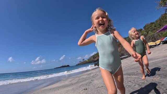 Two happy little kids blonde girls running to camera on summer beach. Dynamic video of smiling girls preschoolers in swimsuit laughing, running races along beautiful beach Running on beach to have fun