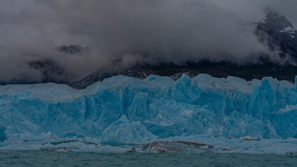 The famous Perito Moreno glacier. A wall of blue cracked ice over a turquoise glacial lake. Thawed ice floes, icebergs float in the water. The mountains are hidden in clouds and fog. El Calafate. 