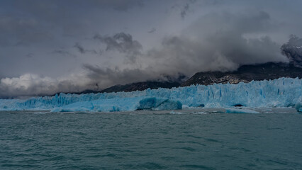 A wall of blue ice with cracks and crevices stretches in a glacial lake. Thawed ice floes, icebergs float in turquoise water. Coastal mountains are hidden in clouds and fog. Perito Moreno. El Calafate