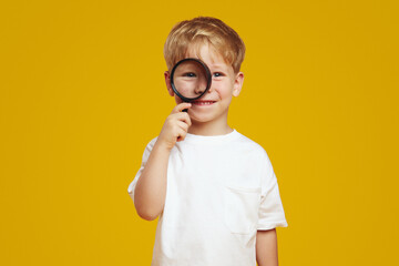 Portrait of charming little child boy with blonde hair wearing white t-shirt, holding magnifier on...