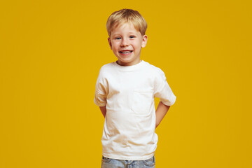 Positive blonde boy in casual white t-shirt keeping hands behind back and smiling for camera while...