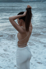 Fototapeta na wymiar Beautiful Asian girl in a white dress posing on the seashore against the backdrop of waves