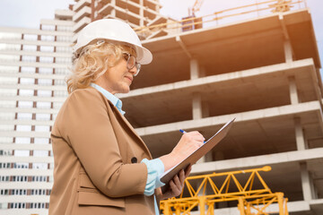 Fototapeta premium Mature businesswoman writes analysis on clipboard at construction site against unfinished building. Blonde architect explores building site, sunlight