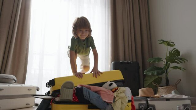Take toys on trip. With reckless abandon, boy jumps on chaotic suitcase, his laughter echoing in room. Randomly arranged items stick out, he's happy with packed travel suitcase. Advertising trip app 