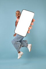 Young joyful ethnic African American woman holds large phone with white screen and jumps up celebrating victory in online game or internet casino posing on isolated turquoise background.