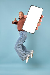 Young excited happy African American woman jumping up and showing off mobile phone and pointing finger at white screen of gadget with copy space for advertising application located in turquoise studio
