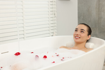 Happy woman taking bath in tub with foam and rose petals indoors