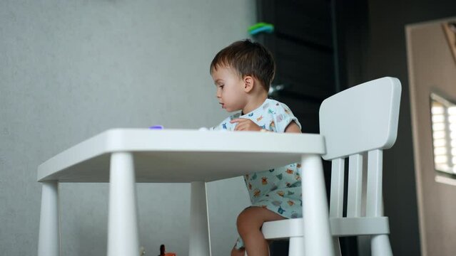 Focused baby boy sits at white desk in his room. Kid plays with his toys cheering and clapping hands.