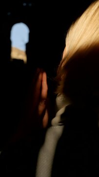 Vertical video: a blonde woman prays in a temple with her palms joined in namaste and asks God for help. The sun's rays shine on a praying woman in a dark temple. faith in God and the power of prayer