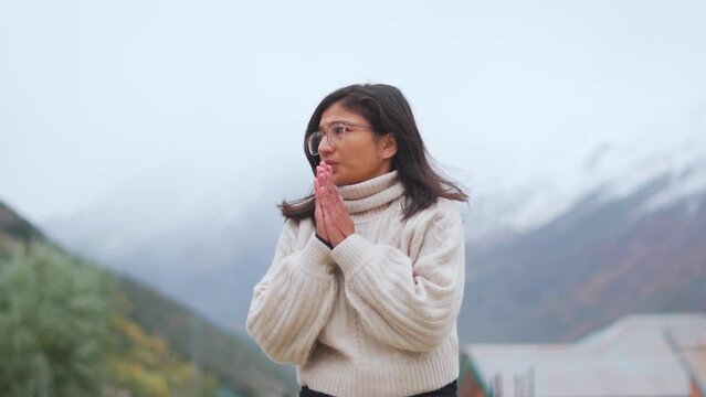 Portrait of Indian teenager girl rubbing hands during cold winter day in Himachal Pradesh, India. Girl wearing warm clothes and standing against snowy Himalayan mountains.