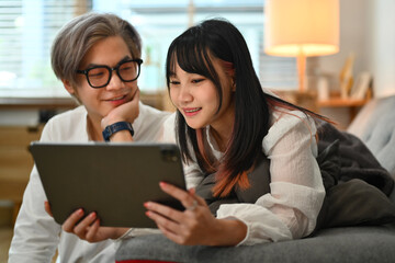 Cheerful young Asian couple sitting on couch and watching movie on digital tablet at home