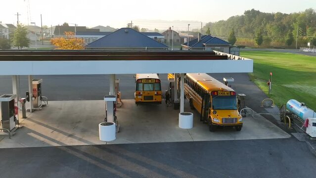 Yellow school buses refueling at small town gas station in America after driving route with students. Aerial establishing shot during sunrise.