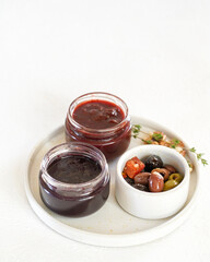 two small jars with red jam and sauce and a bowl with olives on a round wooden board, light background, close-up