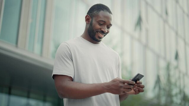 Contented man reading message good news in smartphone typing answer chatting online in messenger social media. Smile african american guy looking at mobile phone screen stand on city street outdoors.